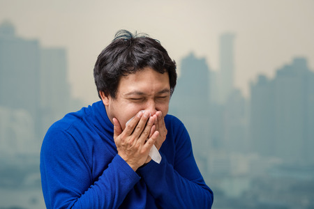 Asian man coughing with Tissue against air pollution with at the balcony of High Apartment which can see pollution and heavy fog over the bangkok cityscape background, healthcare conceptの写真素材