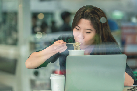 Portrait of asian businesswoman in casual suit eating noodles with happiness action in rush hour at the desk beside the glass in modern office, Business work hard conceptの写真素材