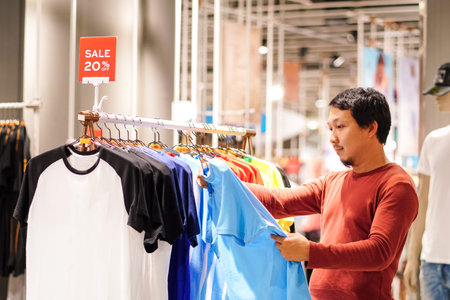 Smart man with beard choosing clothes in clothing store at shopping center, looking t-shirts sale...