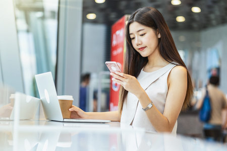 Asian young female holding a cup of coffee and using mobile phone with technology laptop in modern...