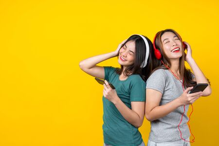 Two Happiness Asian smiling young woman wearing wireless headphones for listening music via smart mobile phone and dancing on isolated yellow color background, Lifestyle and leisure with hobby conceptの写真素材