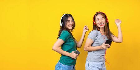 Two Happiness Asian smiling young woman wearing wireless headphones for listening music via smart mobile phone and dancing on isolated yellow color background, Lifestyle and leisure with hobby conceptの写真素材