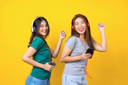 Two Happiness Asian smiling young woman wearing wireless headphones for listening music via smart mobile phone and dancing on isolated yellow color background, Lifestyle and leisure with hobby conceptの写真素材