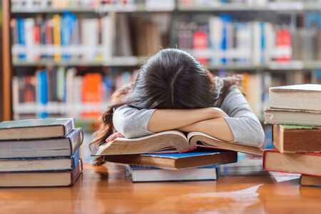Asian young Student in casual suit reading and sleeping on the wooden table with various book in library of university or colleage over the book shelf, Back to school conceptの写真素材