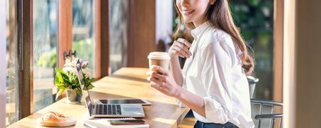 Banner of Portrait Young Asian woman holding and drinking a cup of coffee and working with technology laptop at a coffee shop.freelancer and entrepreneur working by connecting to internet via computerの写真素材
