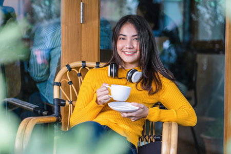 Asian young female holding a cup of coffee and sitting in modern coffee shop or coworking space...