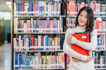 Portrait Asian young Student in casual suit standing and holding the book at book shelf in library of university or college with various book background, Back to school conceptの写真素材