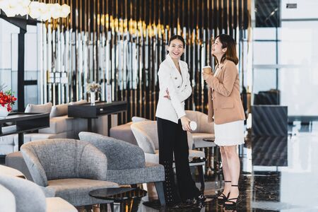 Two asian businesswomen talking during coffee break in modern office or coworking space, coffee break, relaxing and talking after working time, business and people partnership conceptの写真素材