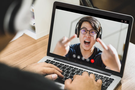 Asian business man wearing headphone and using laptop for video conference with angry colleague showing thumbs up sign Fuck when online meeting, doing fucking off to camera, Social distancing conceptの写真素材
