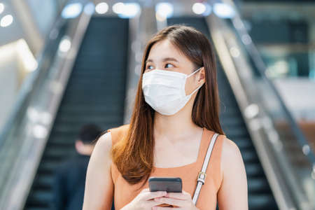 Young Asian woman passenger wearing surgical mask and using smart mobile phone in front of escalator between walk though subway interchange when traveling in big city at Covid19 outbreak, new normalの写真素材