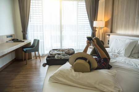 Happiness Asian traveler woman sleeping and using the smart mobile phoneon the bed in bedroom of hotel or hostel when traveling on her holiday,luxury interior bedroom design,tourism and travel conceptの写真素材