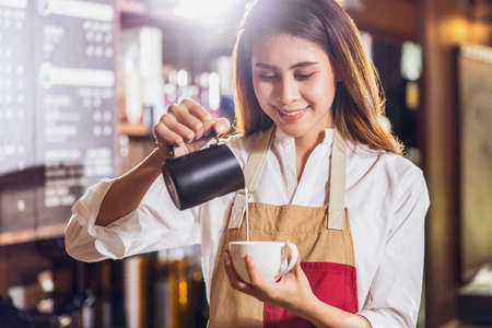 Asian Barista pouring milk to cup of coffee which is espresso with latte or cappuccino for customer order in coffee shop, Small business owner and startup in coffee shop and restaurant conceptの写真素材