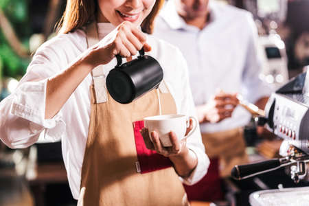 Asian Barista pouring milk to cup of coffee which is espresso with latte or cappuccino for customer order in coffee shop, Small business owner and startup in coffee shop and restaurant conceptの写真素材
