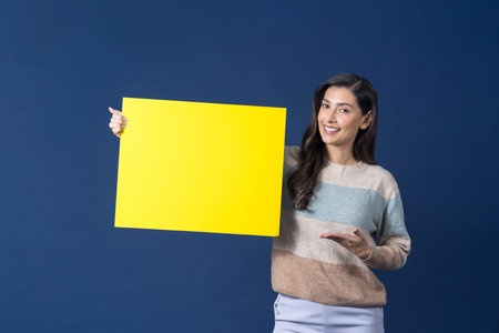 Happy young smiling Asian woman holding and presenting to copy space empty yellow billboard on blue...