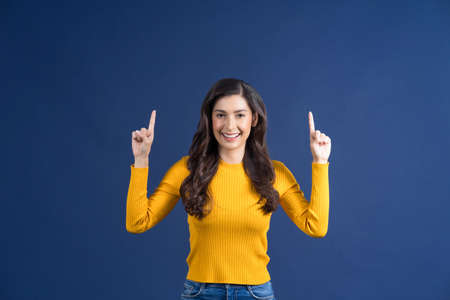 Happy young smiling Asian woman finger Pointing to advertise with happy emotion on blue color background, showing or presenting the product via empty copy space conceptの写真素材