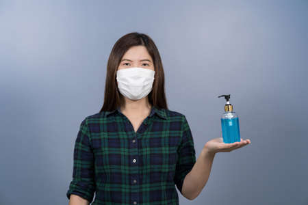 Portrait of asian female wearing face surgical mask and presenting alcohol gel or hand sanitizer on blue color background, covid19 outbreak and pandemic,healthcare and protection against virus conceptの写真素材