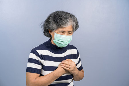 Asian Old woman wearing face surgical mask and  feeling pain ache touching chest having heart attack on blue color background, group risk of covid19 outbreak, protection against coronavirus conceptの写真素材