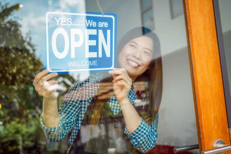 Closeup hand of Asian young woman setting open sign at the shop glasses for welcome the customer in to the coffee shop, small business owner and startup, installing open and close label conceptの写真素材