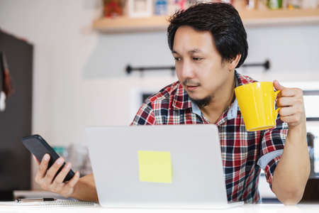 Asian man working from home with technology laptop and mobile phone at kitchen room, Working at home in  pandemic, new normal conceptの写真素材