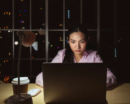 Asian businesswoman working hard late with technology computer in office, customer service and call center, team work with colleagues for success achievement project conceptの写真素材