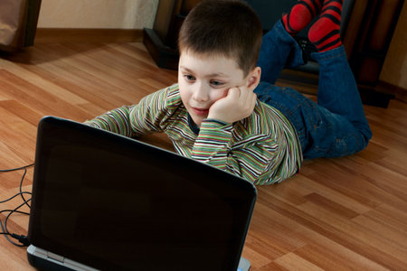 little boy lying down on the floor in the living room and playing computer game on laptop