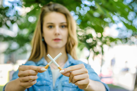 Close Up Of Stylish Smiling Woman Smoking Cigarette Outdoor Denim Shirt in park on Nature Background Habit Prohibited Advertisingの写真素材