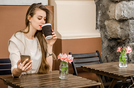 Portrait of a smiling bussines casual woman holding smartphone drinking coffee with long hair in cafe city street sunny day backgroundの写真素材