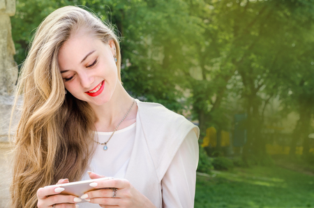 Portrait of a smiling bussines casual woman holding typing text smartphone mobile cell with long hair in cafe city street sunny day backgroundの写真素材