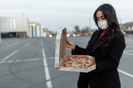 Young woman in medical mask holding cardboard with pizzas going out for food during the epidemic. Concept of takeaway food during quarantineの写真素材