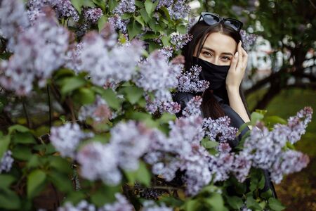 Girl in a medical mask on a background of blooming lilacs. Black mask.の写真素材