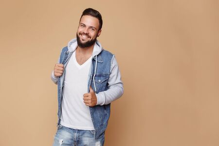 Stylish young cheerful guy with a beautiful beard in a denim jacket and white t-shirt on a plain cream background. Denim jacket advertising concept for billboard.の写真素材
