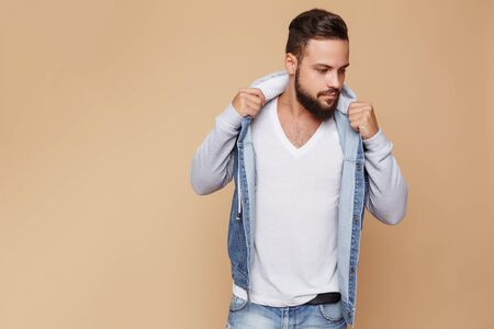 Stylish young cheerful guy with a beautiful beard in a denim jacket and white t-shirt on a plain cream background. Denim jacket advertising concept for billboard.の写真素材