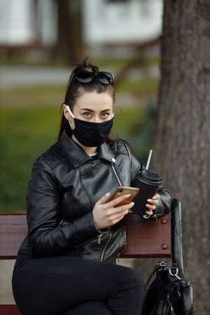 Portrait of brunette woman in a black protective mask sitting on a bench in the park, sunny spring day. Woman talking on mobile phoneの写真素材