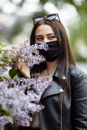 Girl in a medical mask on a background of blooming lilacs. Black mask. Protection against virus, flu. Coronavirus protection. epidemic of coronavirus. the aroma a tree in the garden Spring allergyの写真素材