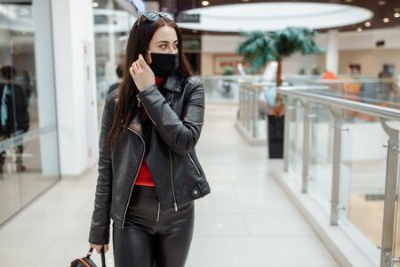 A girl with a medical black mask is walking along a shopping center. Coronavirus pandemic. A girl in a protective mask is shopping at the mallの写真素材