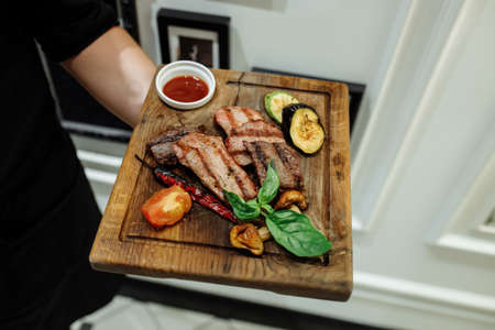 Three types of grilled meat with grilled vegetables on a wooden board. A waiter hand in a glove with a serving board on which grilled meat.の写真素材