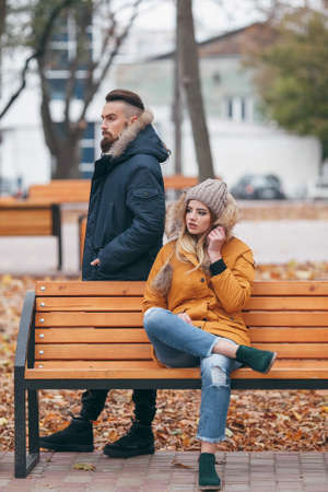 A guy and a girl are resting on a bench in an autumn park. A loving couple in jackets sits on a bench in the main park.の写真素材