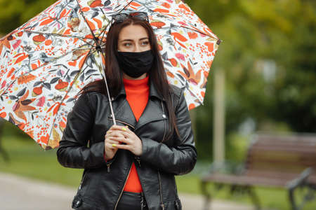 A masked girl is walking along the street. A girl in a protective mask walks in the park with an umbrella in the rain. Coronavirus infection COVID-19.の写真素材