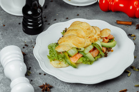 Croissant sandwich with red fish, avocado, fresh vegetables and arugula on black shale board over black stone background. Healthy food concept.の写真素材