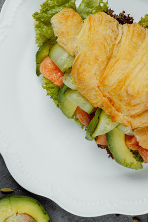 Croissant sandwich with red fish, avocado, fresh vegetables and arugula on black shale board over black stone background. Healthy food concept.の写真素材