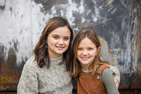 Two young sisters sitting next to each other and hugging with their arms around each other in the winter sitting near an urban warehouse wallの写真素材