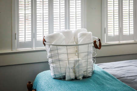A basket of organized clean rolled white towels for guests sitting on a bed in a guest bedroomの写真素材