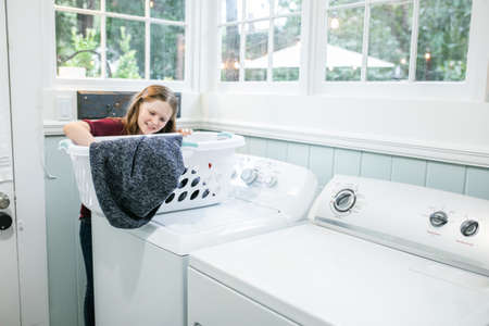 A little girl with long hair and freckles holding a laundry basket and doing the wash as laundry chores in a natural light laundry room with a washer and dryer.の写真素材