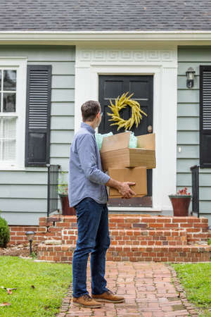 A man and father holding moving boxes outside a small blue cottage house getting ready to move into his new houseの写真素材