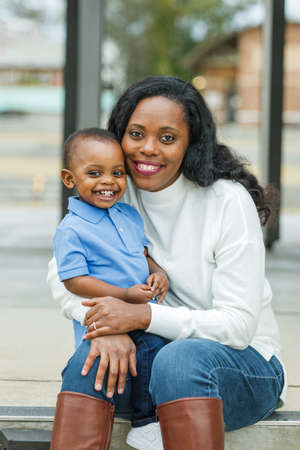 A beautiful African-American mom sitting on steps outdoors and hugging her toddler age son and they are both smiling and happyの写真素材