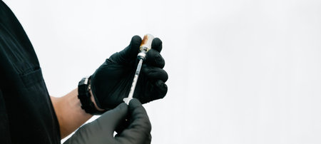 Close up of a medical provider wearing black scrubs and gloves filling a medical syringe against a...