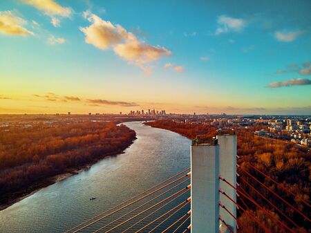 Beautiful panoramic aerial drone view to cable-stayed Siekierkowski Bridge over the Vistula river and Warsaw City skyscrapers, Poland in gold red autumn colors in November evening at sunsetの写真素材