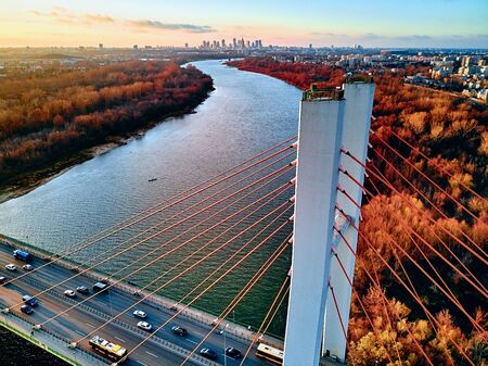 Beautiful panoramic aerial drone view to cable-stayed Siekierkowski Bridge over the Vistula river and Warsaw City skyscrapers, Poland in gold red autumn colors in November evening at sunsetの写真素材