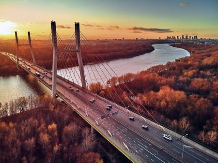 Beautiful panoramic aerial drone view to cable-stayed Siekierkowski Bridge over the Vistula river and Warsaw City skyscrapers, Poland in gold red autumn colors in November evening at sunsetの写真素材
