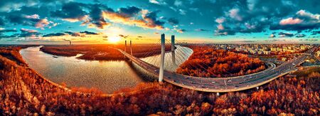Beautiful panoramic aerial drone view to cable-stayed Siekierkowski Bridge over the Vistula river and Warsaw City skyscrapers, Poland in gold red autumn colors in November evening at sunsetの写真素材